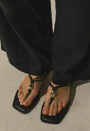 Lábak fekete, négyzetorrú szandált viselnek fekete bőrpántokkal és arany dekoratív díszítésekkel, fekete bőszárú nadrággal párosítva, világos padlón.