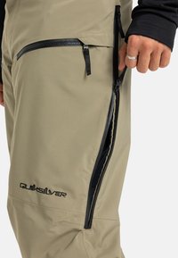 Main dans le processus de dézipper la fermeture éclair de ventilation latérale sur un pantalon d'extérieur marron clair avec un logo Quiksilver noir et des détails de fermeture éclair noirs.