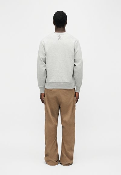 Mann som står med ryggen til iført lys grå genser med lite astronautgrafikk på øvre rygg og løse brune bukser.