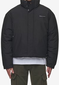 Sort cropped puffjakke med høj krave, lynlåslommer og diskret branding på brystet. Glat mat overflade og polstret tekstur.