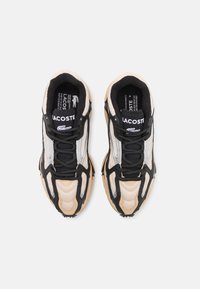 Lacoste sneaker, amely bézs, fekete és ezüst anyagok keverékéből készült. A dizájn hálós panelekből, texturált részletekből és gumitalpakból áll.