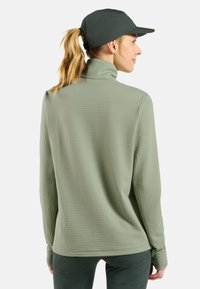 Hellegrünes Langarmoberteil mit einem hohen Kragen, strukturiertem Stoff und Daumenlöchern, kombiniert mit einer dunkelgrünen Mütze.