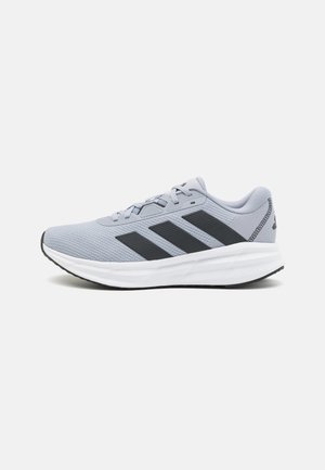 Chaussure de sport gris clair avec des rayures noires, semelle blanche et lacets, vue de côté sur un fond blanc uni.