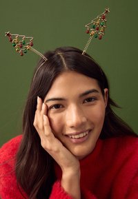Gouden hoofdband met twee accenten in de vorm van kerstbomen, versierd met rode en groene kralen, tegen een groene achtergrond.