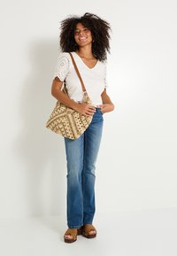 Bolso de mano texturizado beige con detalles trenzados y una correa de hombro marrón, combinado con una blusa clara y unos jeans acampanados azules. Sandalias de plataforma marrones.