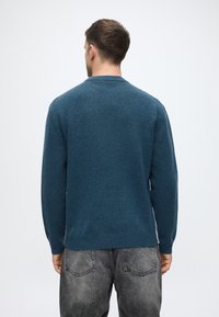 Mīksta adīta džemperis tumši zilā krāsā ar apaļu kakla izgriezumu un ribotām piedurknēm. Nēsā virs pelēkajām nolietotajām džinsām, ar gludu tekstūru.