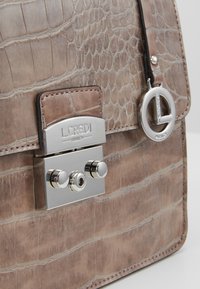 Beige handtas met krokodillenprint en zilverkleurige hardware, voorzien van een druksluiting en een ronde logo-bedel aan het handvat.