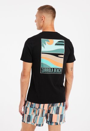 Sort bomulds T-shirt med et grafisk design på bagsiden, der viser abstrakte strandscener i teal, orange og beige. Model iført mønstrede badebukser.