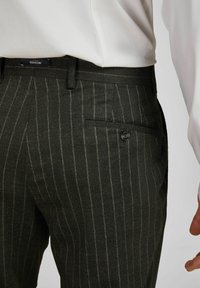 Dunkelgrüne Hose mit einem vertikalen Nadelstreifenmuster, ausgestattet mit einer Knopftasche auf der Rückseite und einem flachen Vorderdesign. Hergestellt aus einem strukturierten Stoff.