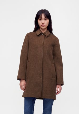 Femme portant un manteau marron à carreaux, longueur genoux, avec col, fermeture par boutons, et un jean bleu sur un fond blanc uni.