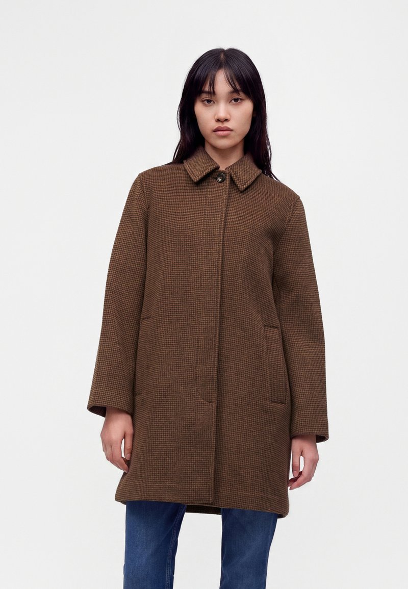 Femme portant un manteau marron à carreaux, longueur genoux, avec col, fermeture par boutons, et un jean bleu sur un fond blanc uni.