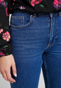 Mörkblå denimjeans har en slät yta och en klassisk femficksdesign. En blus med blommönster och rynkade ärmar är delvis synlig.
