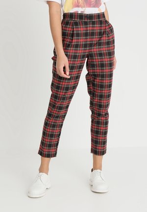 Vrouw draagt rood-zwart geruite, hoog getailleerde taps toelopende broek met zakken en witte veterschoenen, staand tegen een effen achtergrond.