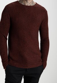 Red textured long-sleeve sweater with a ribbed design and a round neckline, paired with dark pants, highlighting the fabric's knit pattern.