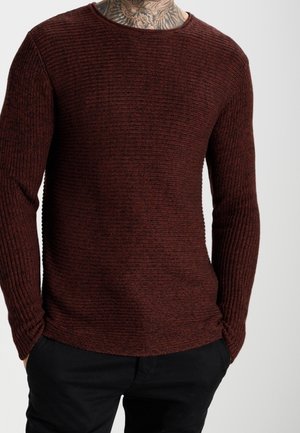 Pull long à manches longues texturé rouge avec un design côtelé et un col rond, associé à un pantalon sombre, mettant en valeur le motif tricoté du tissu.