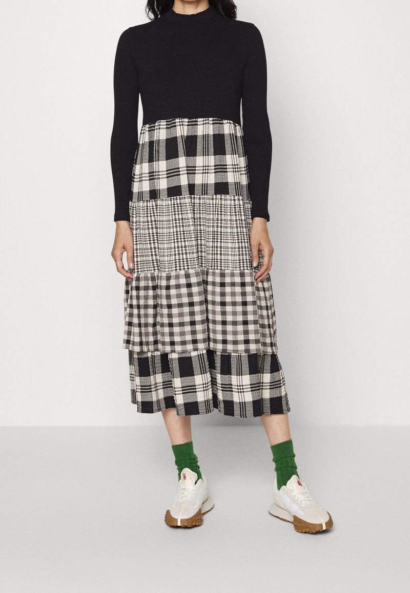 Femme portant un haut noir à manches longues et une jupe midi à volants avec des motifs à carreaux noirs et blancs mélangés, des chaussettes vertes et des baskets blanches.