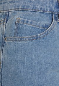 Tessuto di denim azzurro chiaro con una superficie leggermente testurizzata, dotato di una tasca frontale con una linea di cucitura visibile e un dettaglio in rivetto metallico.