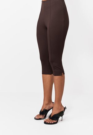 Brązowe, krótkie, dopasowane spodnie z przednim szwem i małymi bocznymi rozcięciami, noszone z czarnymi sandałami na wysokim obcasie z piórowymi detalami.