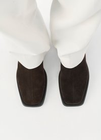 Scarpe in suede marrone con una punta quadrata, caratterizzate da una texture liscia e una suola in gomma nera. Indossate con pantaloni bianchi.