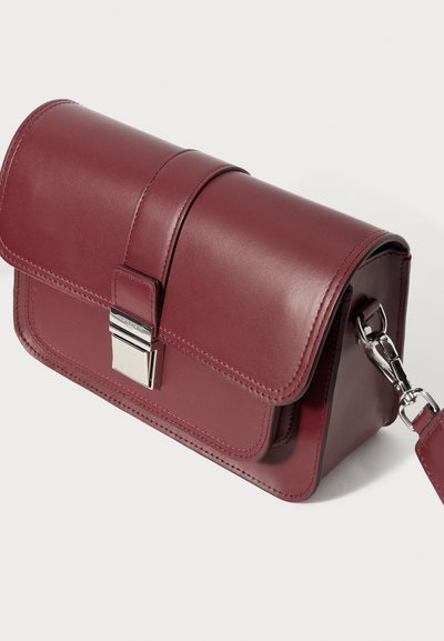 Τσάντα χειρός από δέρμα μπορντό με κλείσιμο με καπάκι, ασημένιες μεταλλικές λεπτομέρειες και λουρί-αλυσίδα. Διαθέτει ραφές διακόσμησης και ορθογώνιο σχήμα.