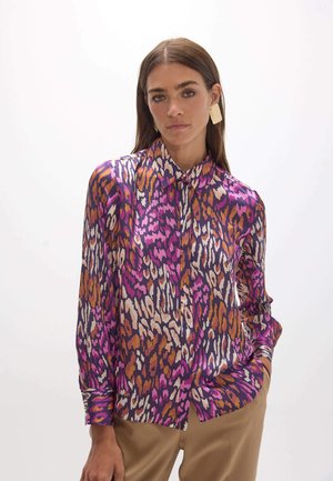 Mujer que lleva una blusa de botones de manga larga con un estampado abstracto de leopardo en rosa, morado, naranja y blanco, combinada con pantalones beige.