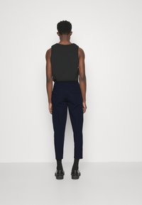 Svart ärmlös topp som kombineras med marinblå tapered byxor med två bakfickor. Modellen bär svarta strumpor och skor, sedd bakifrån.