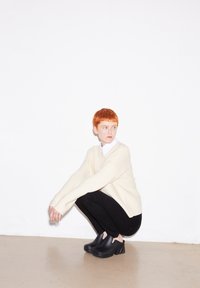 Schwarze Clogs mit einem stabilen Absatz, getragen mit schwarzen Hosen und einem cremefarbenen Pullover mit langen Ärmeln, vor einem weißen Hintergrund.