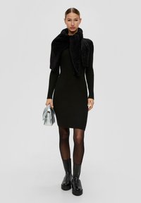 Schwarzes, geripptes Kleid mit langen Ärmeln, kombiniert mit einem strukturierten schwarzen Schal und einer glänzenden silbernen Clutch. Das Model trägt transparente schwarze Strumpfhosen und klobige schwarze Stiefel.