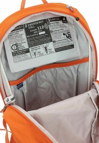 Interno di uno zaino arancione con fodera bianca, caratterizzato da un pannello stampato con istruzioni di emergenza e numerosi scomparti per l'organizzazione.