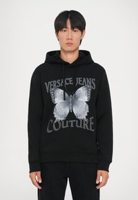 Hanorac din bumbac negru, cu un grafic mare de fluture gri și textul "VERSACE JEANS COUTURE". Proiectat cu un glugă cu șnur și manșete ribbed.