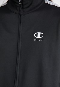 Casaco preto com fecho à frente e gola alta, feito de tecido suave. Apresenta o logótipo branco "Champion" no peito e riscas brancas nos ombros.