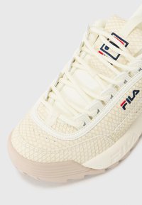Kreemivärvi sneaker, millel on nahkhiirte tekstuur, ümar varvas, paks kummist tallapind ja Fila logo tumesinise ja punase värviga külje peal.