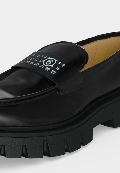 Μαύρο δερμάτινο loafer με χοντρή σόλα και λευκούς τυπωμένους αριθμούς και έναν κύκλο με το νούμερο έξι στο πάνω λουρί.