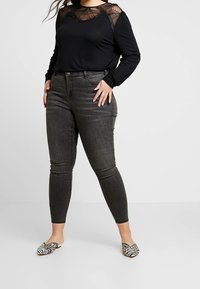 Schwarzes Langarmshirt mit Spitzenverzierungen an den Schultern, kombiniert mit dunkelgrauen Skinny-Jeans und gemusterten Slippern.