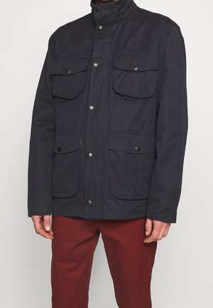 Mann trägt eine schwarze, geknöpfte Jacke mit vier vorderen Klappentaschen und eine rote Hose vor einem einfarbigen Hintergrund.