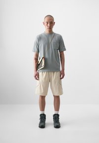 T-shirt gris à manches courtes avec une poche sur la poitrine, shorts beiges et bottes foncées. Un sac à bandoulière pend à l'épaule, avec une couleur contrastante.
