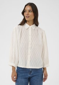 Blouse blanche boutonnée avec un motif zigzag texturé, manches longues bouffantes et col classique. Associée à un jean en denim bleu.