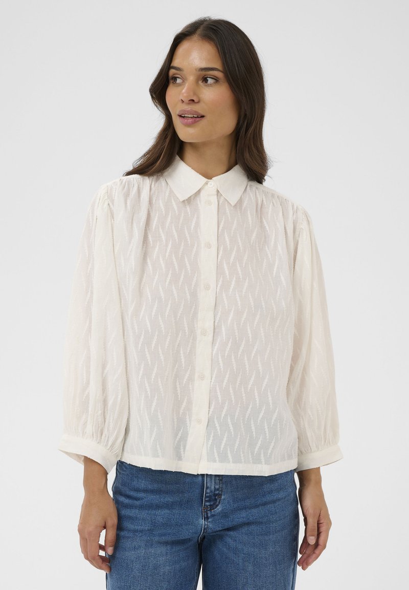 Blouse blanche boutonnée avec un motif zigzag texturé, manches longues bouffantes et col classique. Associée à un jean en denim bleu.