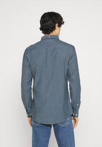 Pánská košile s dlouhým rukávem v tmavě modré látce, s límečkem na knoflíky, zády s plisováním a nastavitelnými manžetami. Hladká textura a střih na míru.