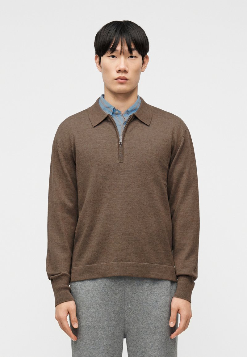 Νεαρός άνδρας με μαύρα μαλλιά, φοράει καφέ πουλόβερ με φερμουάρ πάνω από μπλε πουκάμισο και ανοιχτό γκρι παντελόνι, στέκεται μπροστά σε λευκό, απλό φόντο.