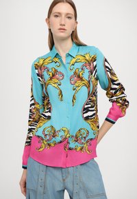 Blusa dai colori vivaci con un corpo turchese, accenti rosa e intricati motivi floreali dorati e rosa. Maniche lunghe con lati a righe.