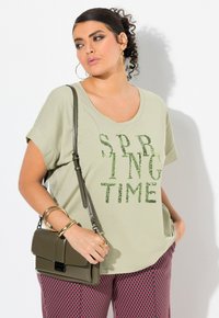 Camiseta suelta de color verde claro con "SPRING TIME" en texto de lentejuelas verdes. Combinada con pantalones de cuadro rosa y un bolso cruzado de color verde oliva.