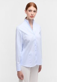 Lichtblauwe shirt met een staande kraag, knoopsluiting aan de voorkant, lange mouwen en een subtiel textuurpatroon op de stof.
