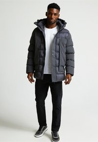 Uomo che indossa un giubbotto imbottito grigio scuro con cappuccio, una maglietta grafica bianca, pantaloni neri e sneaker grigie mentre si trova su uno sfondo neutro.