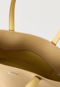 Μπεζ υφασμάτινη τσάντα ώμου με κίτρινη εσωτερική επένδυση και χερούλια, με ανοιχτό πάνω μέρος και εσωτερική τσέπη με φερμουάρ, σε λευκό φόντο.