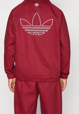 Mann trägt einen roten Trainingsanzug mit einem weißen, umrissenen Trefoil-Logo auf der Rückseite der Jacke, steht vor einem einfarbigen Hintergrund.