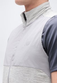 Približni posnetek osebe, ki nosi svetlo sivo brezrokavnik čez temno sivo majico z visokim ovratnikom in sprednjo zadrgo.