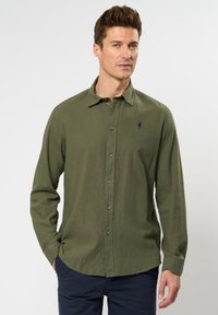 Maslinasto zeleni dugosleeve majica s gumbima, ima gumbasti ovratnik, gumbasti placket, i mali mornarski plavi logo na prsima; izrađena je od teksturirane tkanine.