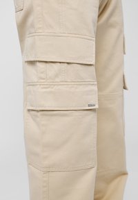 Pantalon cargo beige en tissu de coton résistant, doté de multiples poches latérales, de coutures renforcées et d'une petite étiquette de marque sur la jambe.