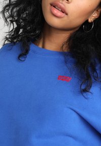 Niebieska bawełniana bluza z kapturem z czerwonym logo Levi's na górnej lewej stronie. Tekstura jest miękka, a dekolt jest ściągaczowy.